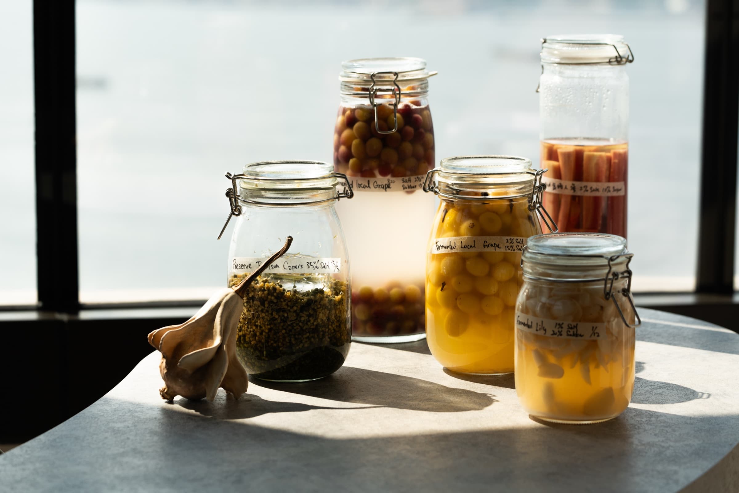 Glass jars of pickled grapes, capers, rhubarb, and pickled lily bulb on a table with a dried garlic bulb, Victoria Harbour softly visible through the window behind.
