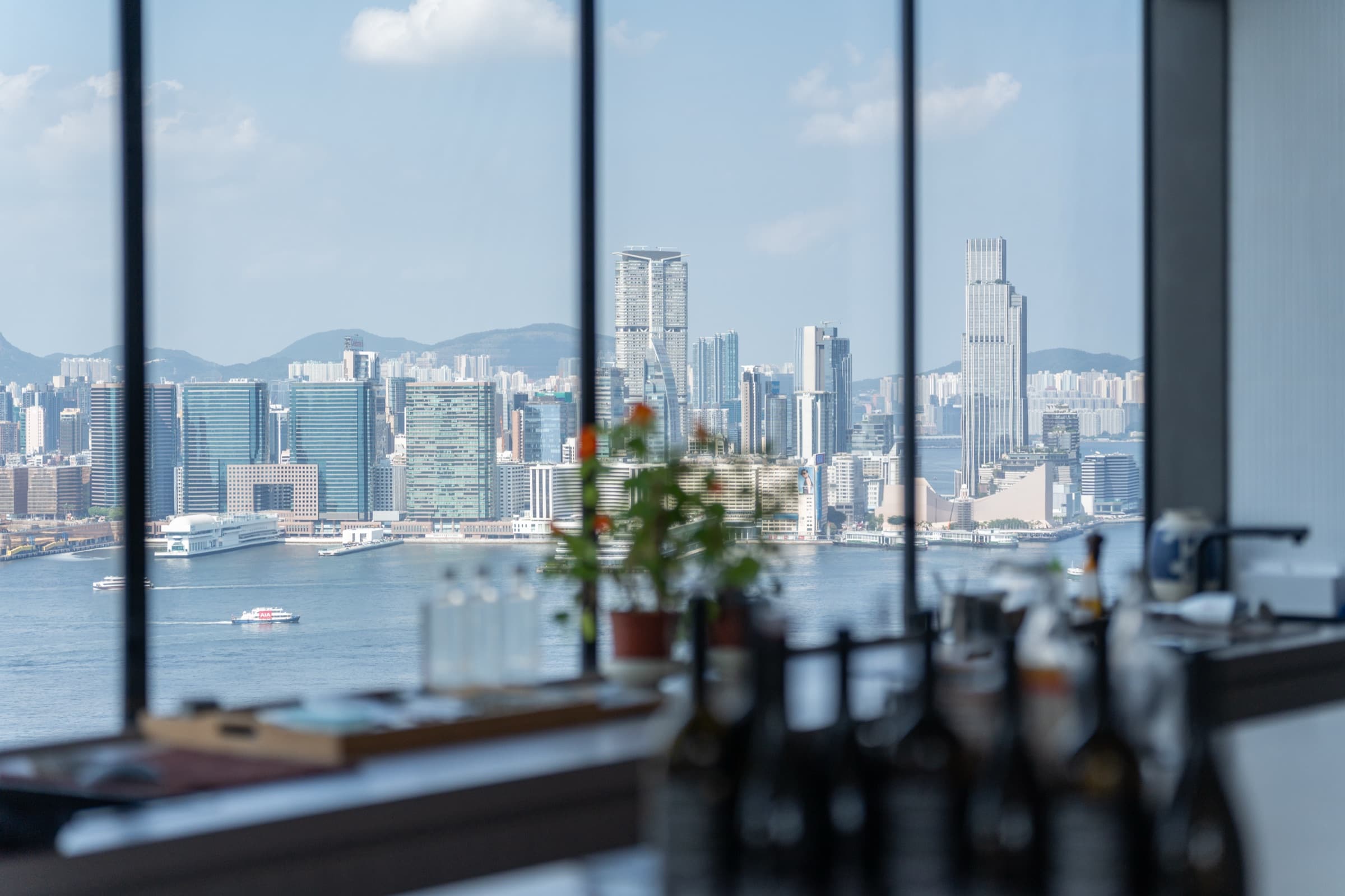 Hong Kong and Kowloon skyline seen through æra's floor-to-ceiling windows, with Victoria Harbour and mountains in the distance.