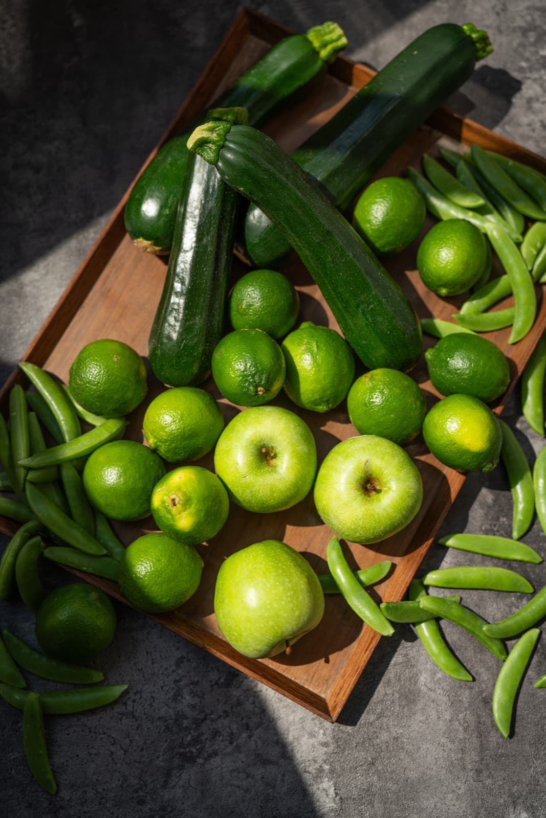 A wooden tray of fresh local produce — zucchini, limes, green apples, and snap peas — arranged in natural light.